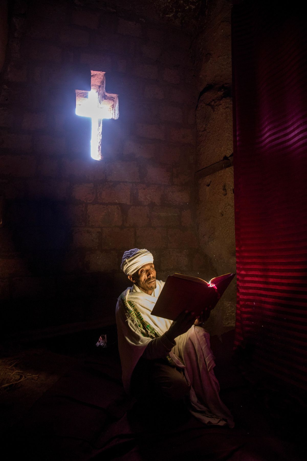A Lalibela priest reads his bible by the light shining through a crucifix shaped 'window' in a rock church
