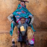 Celebrating the feminine divinity, a young child sits with a masked Guèlèdè dancer in Benin (image by Inger Vandyke)