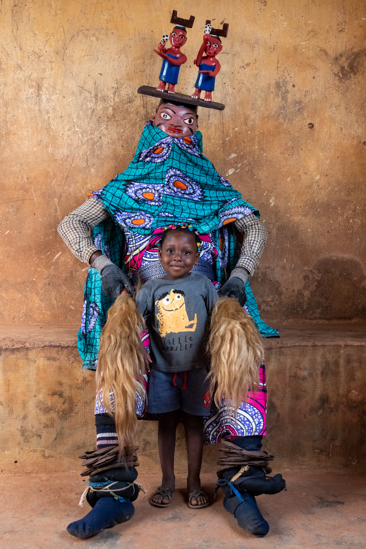 Celebrating the feminine divinity, a young child sits with a masked Guèlèdè dancer in Benin (image by Inger Vandyke)