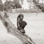 Sepia image of a smiling young Arbore girl on the trunk of an ancient, gnarled tree (image by Inger Vandyke)