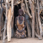 Looking like a jewel in a handmade wall of wood, a Hamer woman sits in the doorway of her temporary agricultural home (image by Inger Vandyke)