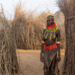 A stunningly decorated Nyangatom stands in the equally stunning homes of her village (image by Inger Vandyke)
