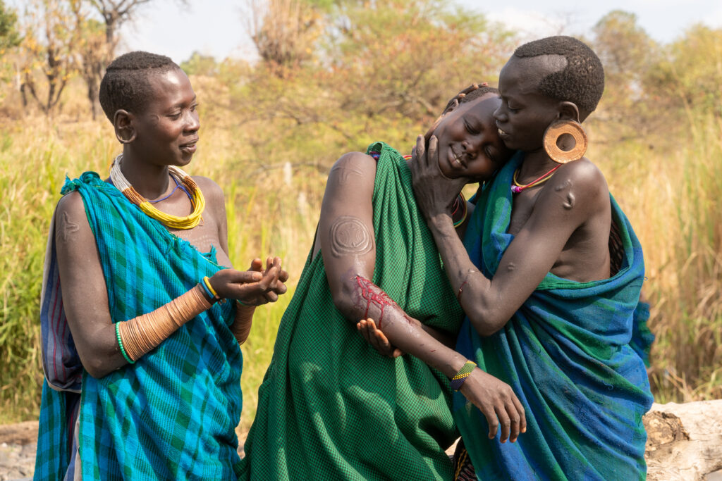 At the end of her scarification, a young Suri girl is supported by her friend and smiles with relief that its over (image by Inger Vandyke)