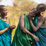 At the end of her scarification, a young Suri girl is supported by her friend and smiles with relief that its over (image by Inger Vandyke)