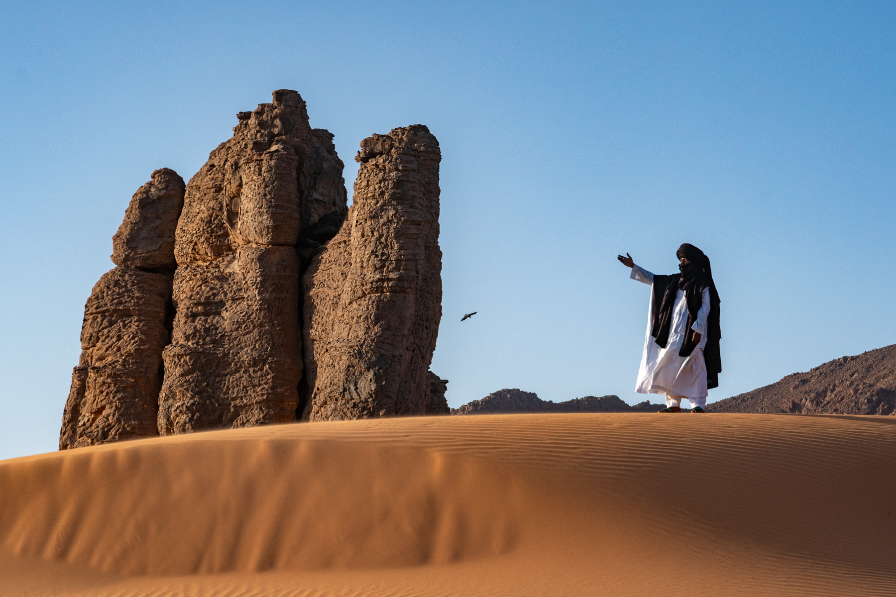 A Tuareg man waves his hand into the air setting a raven into flight in the desert (image by Inger Vandyke)