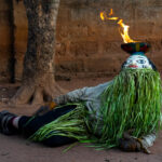 A Guelede Fire Dancer lies on the ground while balancing a bowl of fire on his head! (image by Inger Vandyke)