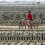 A young boy rides his red bicycle through lines of drying bricks (image by Inger Vandyke)