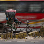 A bus speeds past a homeless cycle rickshaw driver lying asleep on a bed propped up by bricks (image by Inger Vandyke)