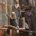 A line of workers chipping rust off the hull of a ship (image by Inger Vandyke)