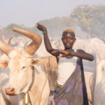 A Mundari girl holds her arms in the air to pose like her favourite cow (image by Inger Vandyke)