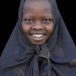 An Arbore girl with a shaved head and a bright smile, colorful beads hidden beneath her scarf. (image by Ingrid Koedood)