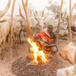 A Mundari man warms himself by a fire at dusk (image by Inger Vandyke)