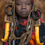 A Mursi girl framed by rich earth tones and vibrant red, her adornments glowing in the warm light. (image by Ingrid Koedood)