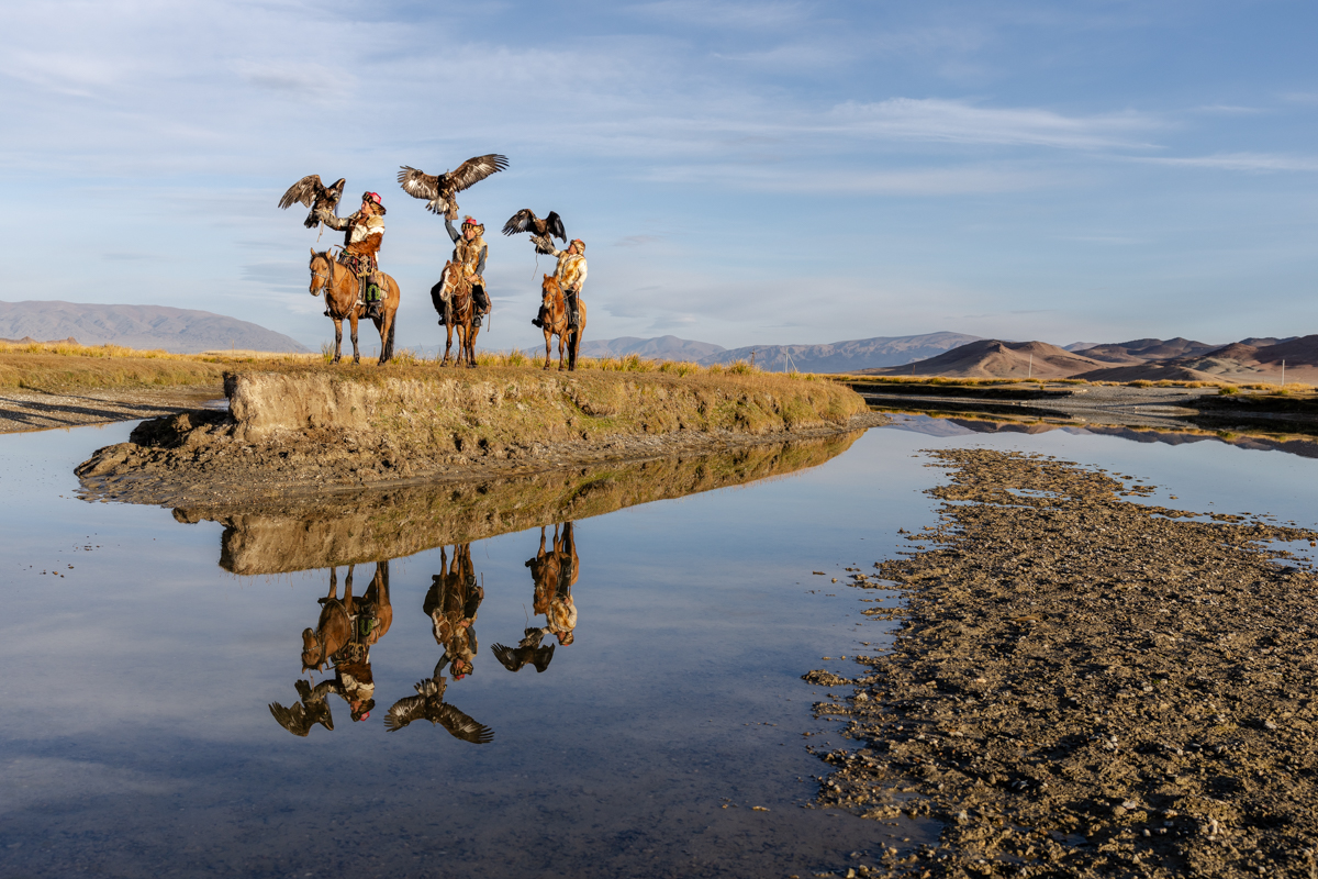Three eagle hunters on horseback with their golden eagles, reflected in the still water during an early morning shoot (image by Ingrid Koedood)